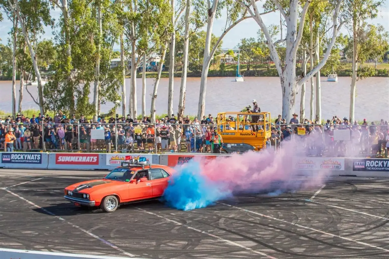 A car doing a burnout in blue and pink smoke, in front of a riverside crowd